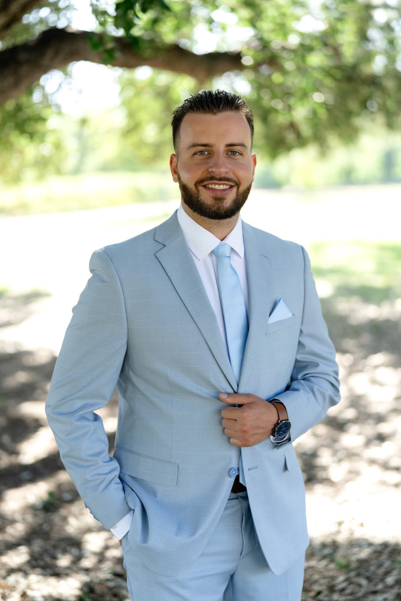 Housam Dandouch in a light blue suit, standing outdoors under a tree